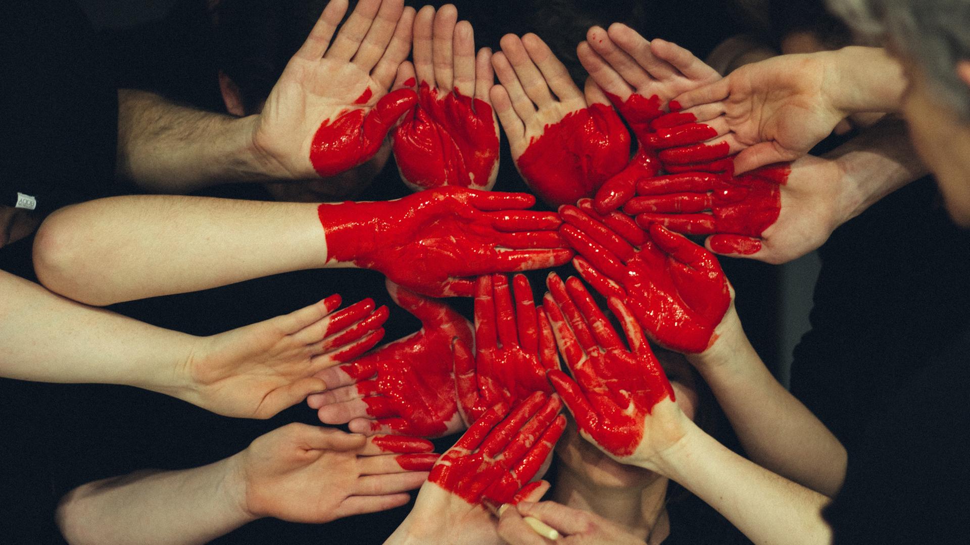 Viele Hände formen eine Fläche, auf die mit roter Farbe ein Herz gemalt ist.
