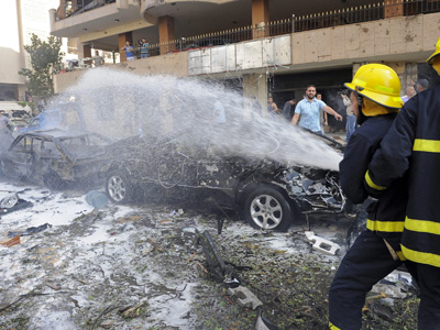 Links im Bild ist ein ausgebranntes Auto zu sehen, recchts ein Feuerwehrmann, der mit einem Wasserschlauch versucht zu löschen..