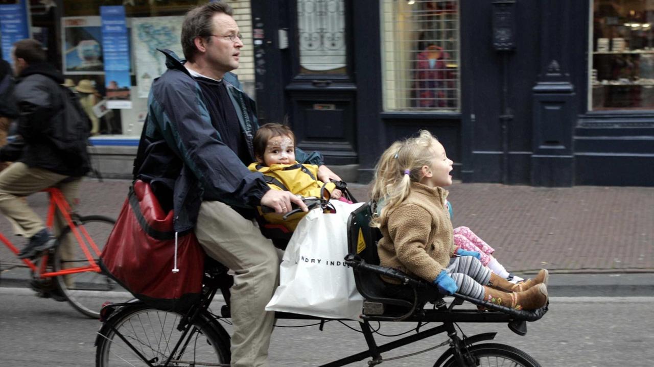 Auf einem ungewöhnlichen Zweirad radelt ein Mann mit seinen beiden Kindern, die in Kindersitzen vorne sitzen, durch die Innenstadt von Amsterdam. Auf einem ungewöhnlichen Zweirad radelt ein Mann mit seinen beiden Kindern, die in Kindersitzen vorne sitzen, durch die Innenstadt von Amsterdam.