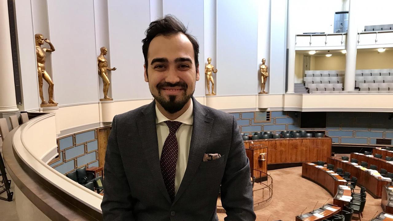Der grüne Abgeordnete im finnischen Parlament, Ozan Yanar, sitzt im ersten Stock des Parlaments auf der Ballustrade. Unten im Hintergrund ist der Plenarsaal zu sehen. Der grüne Abgeordnete im finnischen Parlament, Ozan Yanar, sitzt im ersten Stock des Parlaments auf der Ballustrade. Unten im Hintergrund ist der Plenarsaal zu sehen.