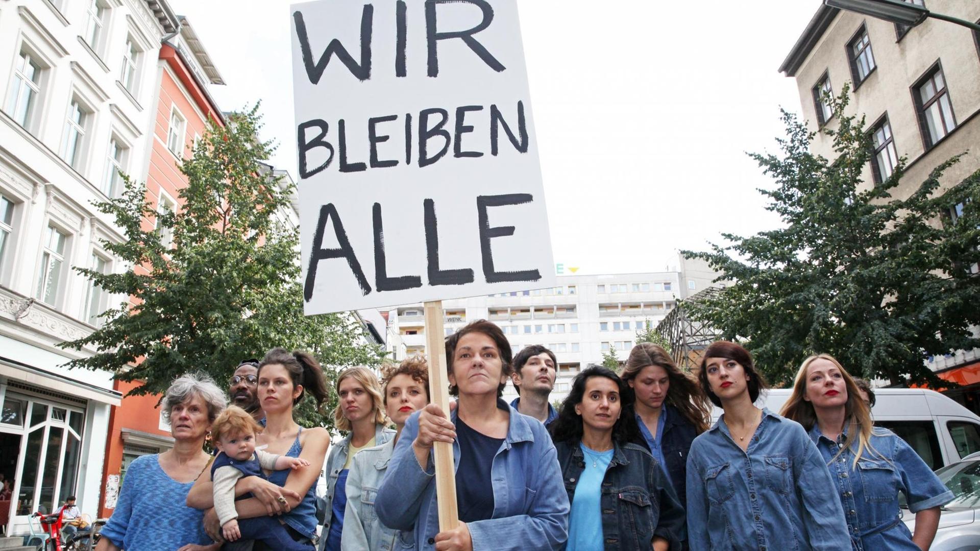 Die Autorin Christiane Rösinger ist mit ihrem Cast zu sehen, sie hält ein Schild mit der Aufschrift "Wir bleiben Alle" hoch.