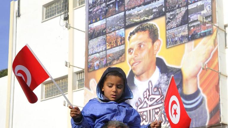 Ein kleines Mädchen sitzt auf den Schultern eines Mannes und hat in beiden Händen kleine tunesische Flaggen. Im Hintergrund ist an einer Hauswand ein großes Porträt von Mohamed Bouazizi zu sehen. Ein kleines Mädchen sitzt auf den Schultern eines Mannes und hat in beiden Händen kleine tunesische Flaggen. Im Hintergrund ist an einer Hauswand ein großes Porträt von Mohamed Bouazizi zu sehen.