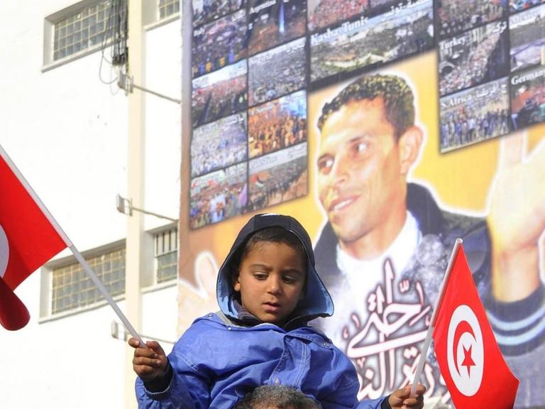 Ein kleines Mädchen sitzt auf den Schultern eines Mannes und hat in beiden Händen kleine tunesische Flaggen. Im Hintergrund ist an einer Hauswand ein großes Porträt von Mohamed Bouazizi zu sehen.