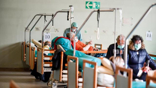 Patienten mit Mundschutzmasken liegen in einem großen Raum aufgereiht in Krankenbetten.
