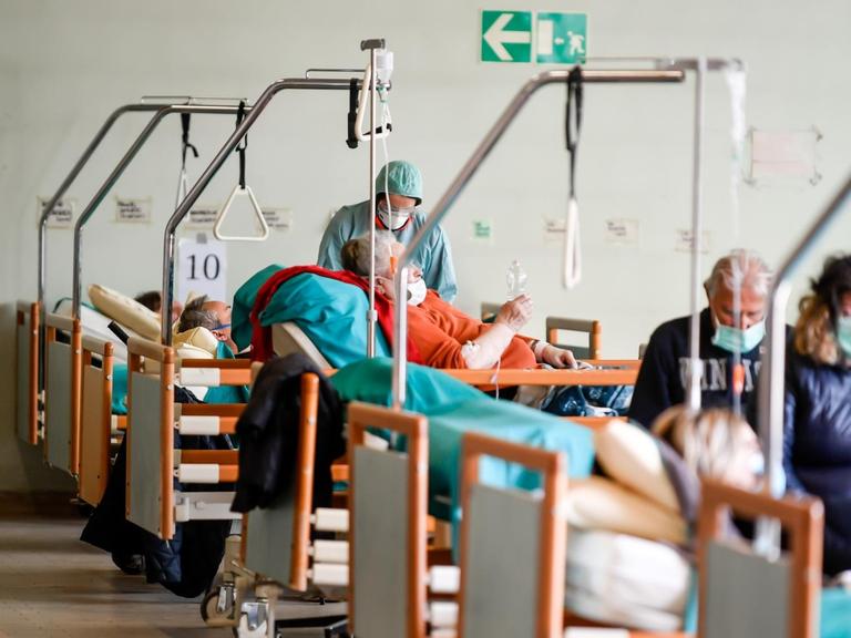 Patienten mit Mundschutzmasken liegen in einem großen Raum aufgereiht in Krankenbetten.