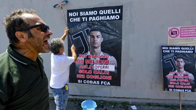 Mitarbeiter von Fiat Chrysler protestieren vor dem Werk in Pomigliano. Auf dem Plakat an den Wänden der ehemaligen Fiat-Fabrik hängt ein Foto des Fußballstars Ronaldo mit der Aufschrift "Wir beschuldigen sie, 400 Millionen Euro auszugeben für einen Fußballspieler und dafür Gewinne zu benutzen, die die Arbeiter dieser Fabrik erwirtschaften."