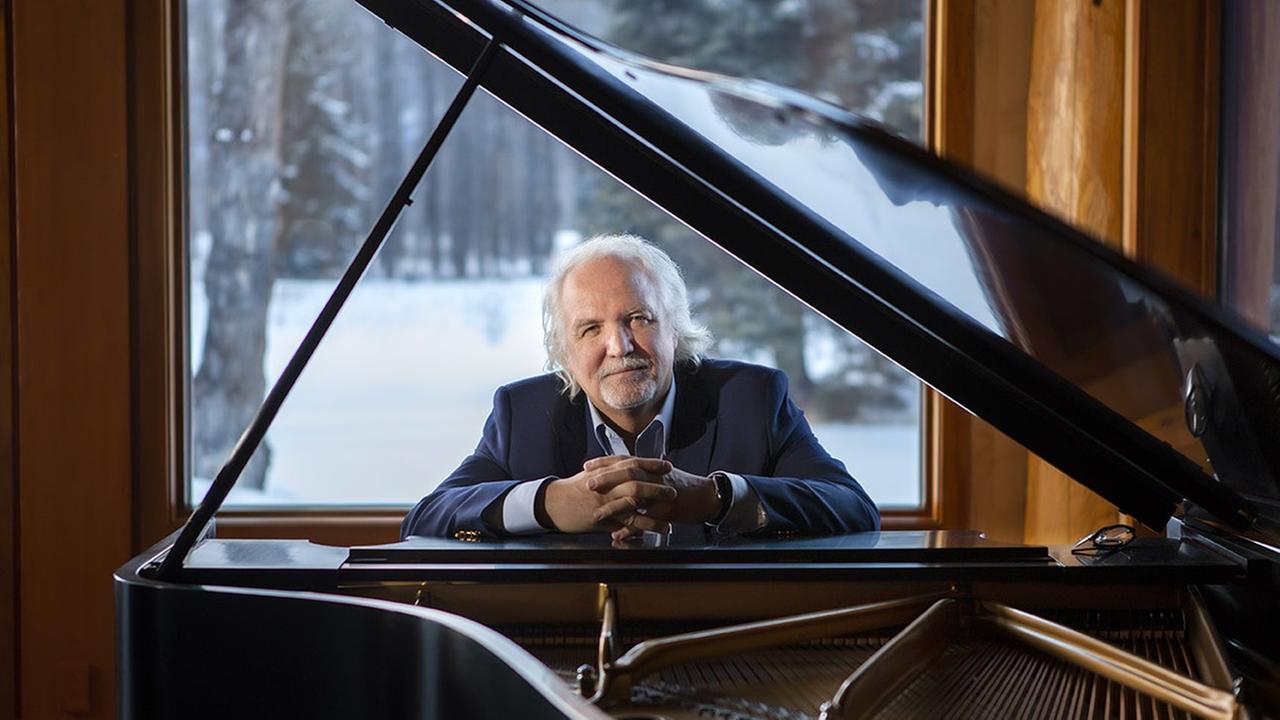 Der Dirigent Donald Runnicles sitzt hinter seinem Piano vor einem Fenster, durch das eine winterliche Landschaft zu sehen ist.