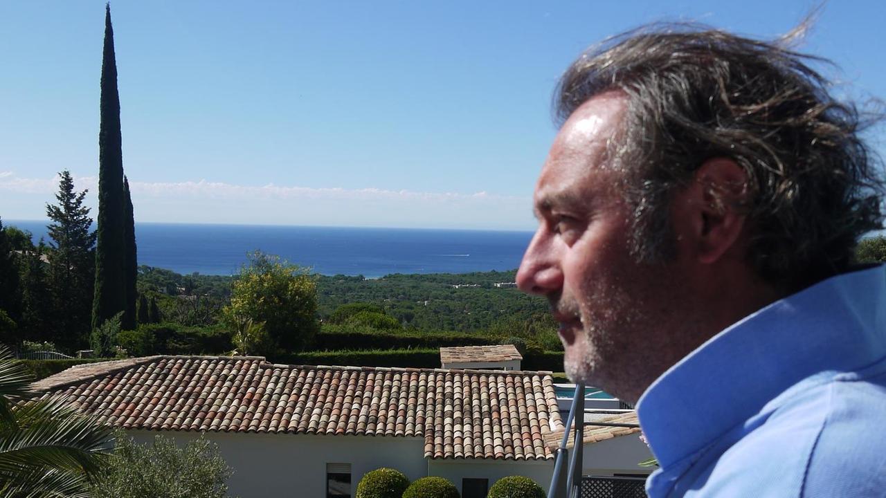 Christoph Metzger im Profil, Balkon, Haus im Süden, hellblaues T-Shirt, Zypresse, blauer Himmel, Meer
