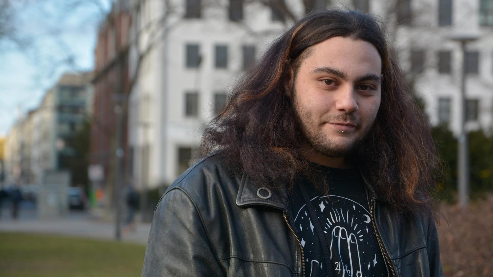 Der Dichter Sam Zamrik trägt eine schwarze Lederjacke und hat langes dunkles Haar. Er steht in einem Park. Im Hintergrund sind Häuser zu erkennen. Der Dichter Sam Zamrik trägt eine schwarze Lederjacke und hat langes dunkles Haar. Er steht in einem Park. Im Hintergrund sind Häuser zu erkennen.