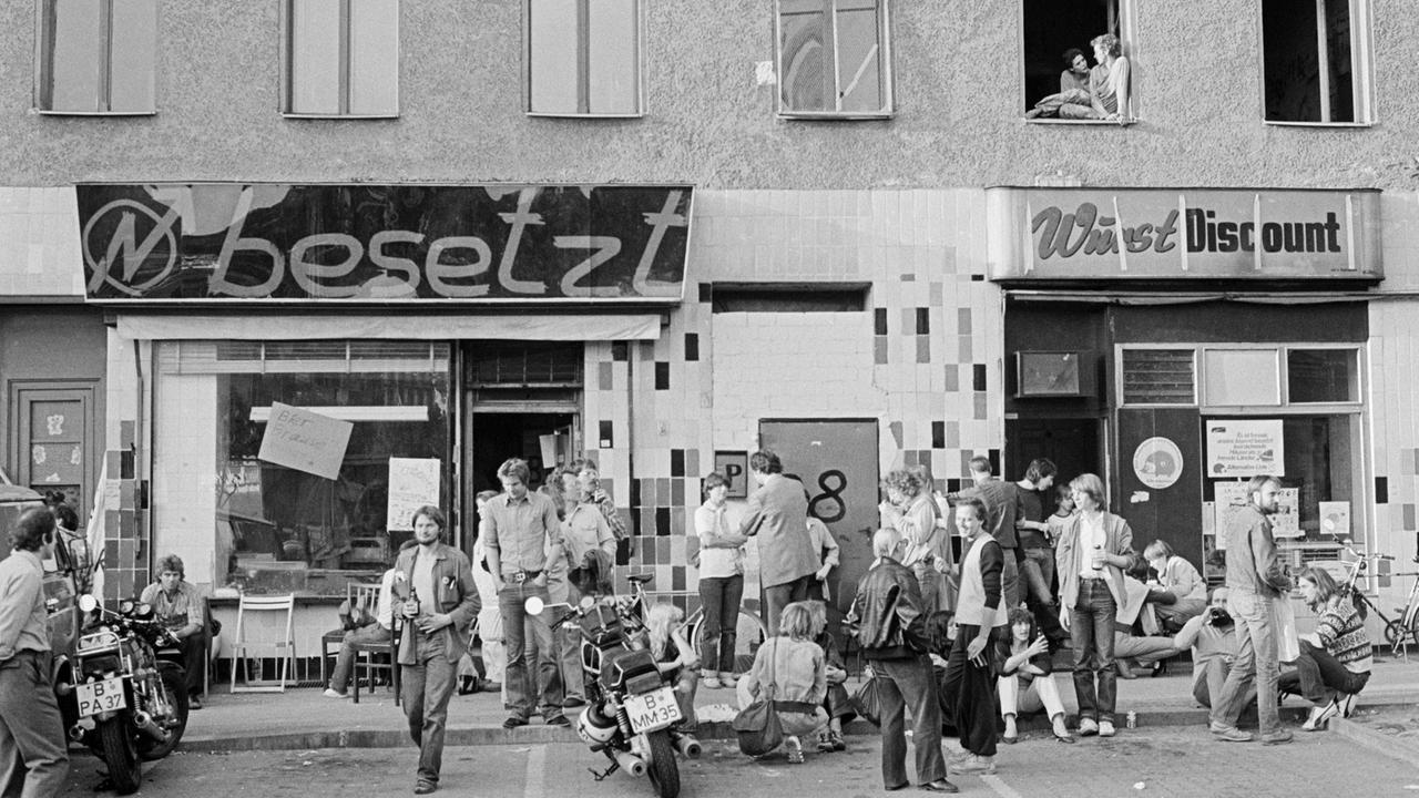 Schwarz-Weiß-Bild einer Hausbesetzung in Kreuzberg in den 80er-Jahren. Menschen sitzen und stehen vor einem besetzten Haus.
