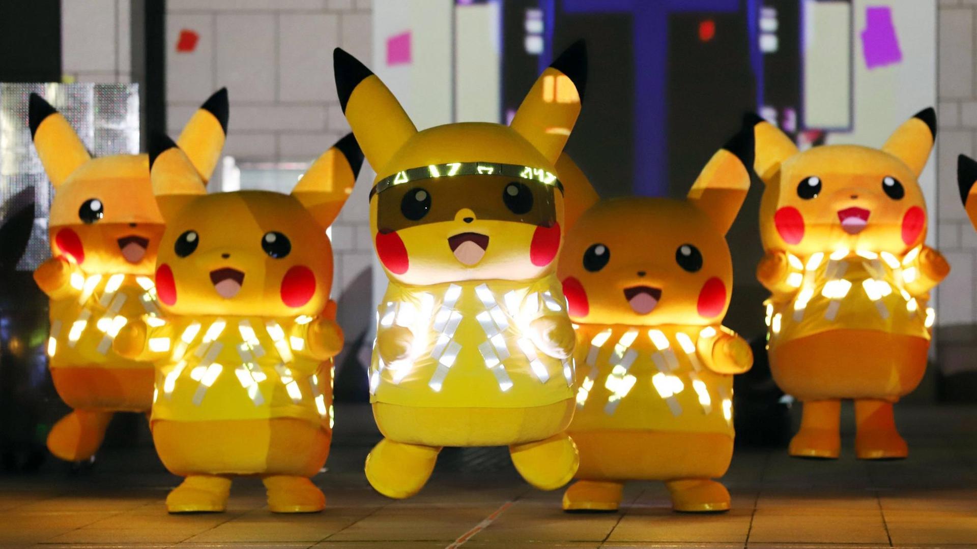 Fünf menschengroße Pikachus (ein gelbes. hasenähnliches Wesen mit langen Ohren, die an der Spitze schwarz sind) mit LED-Lichtern tanzen auf einer Bühne.