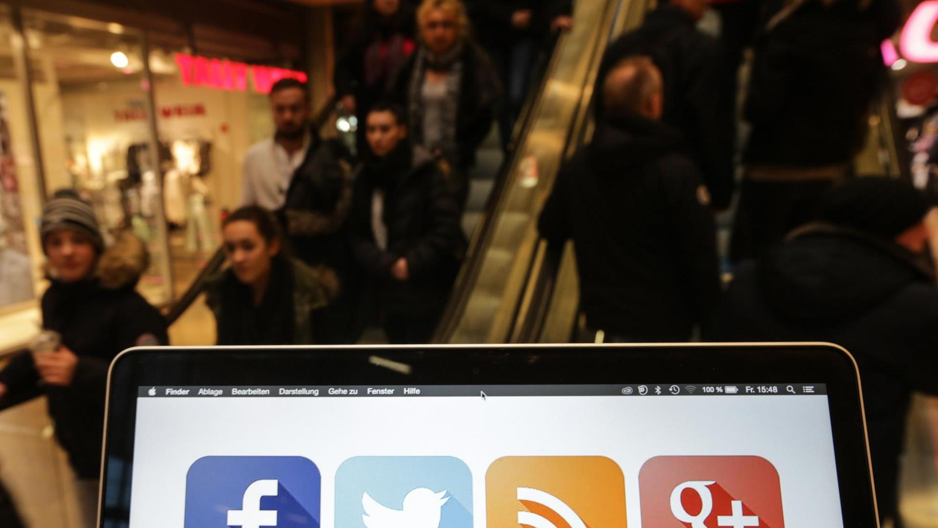 Ein Laptop mit Social Media Icons auf dem Bildschirm, dahinter Menschen auf der Rollstreppe eines Einkaufszentrums in Hamburg. Ein Laptop mit Social Media Icons auf dem Bildschirm, dahinter Menschen auf der Rollstreppe eines Einkaufszentrums in Hamburg.