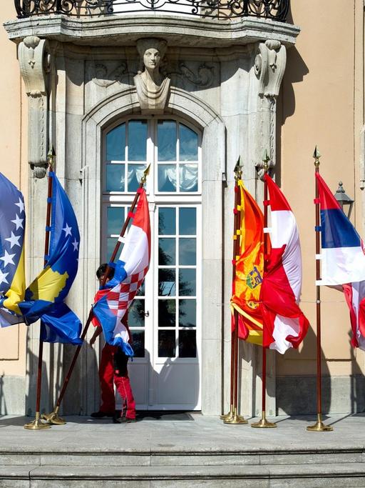 Mitarbeiter des Auswärtigen Amts mühen sich bei der Westbalkan-Konferenz in Berlin vor dem Gruppenbild der Außenminister mit den wehenden Fahnen, die umzukippen drohen.