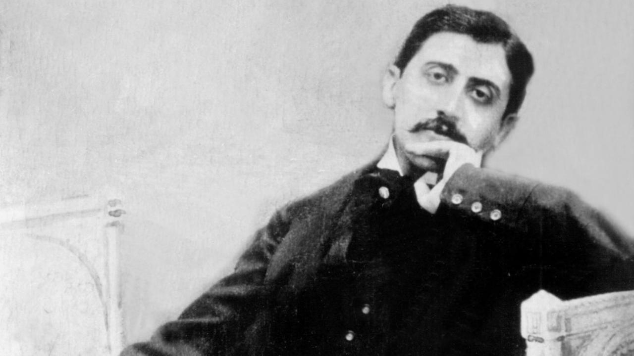 Ein schwarz-weiß Bild von Marcel Proust, der auf einem kleinen Sofa sitzt, mit schwarzen Anzug die linke Hand zur Denkerpose ans Kien gehalten. Er schaut in die Kamera und trägt einen Schnauzer. Ein schwarz-weiß Bild von Marcel Proust, der auf einem kleinen Sofa sitzt, mit schwarzen Anzug die linke Hand zur Denkerpose ans Kien gehalten. Er schaut in die Kamera und trägt einen Schnauzer.