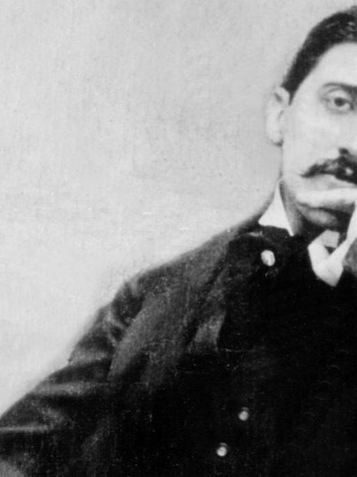 Ein schwarz-weiß Bild von Marcel Proust, der auf einem kleinen Sofa sitzt, mit schwarzen Anzug die linke Hand zur Denkerpose ans Kien gehalten. Er schaut in die Kamera und trägt einen Schnauzer.