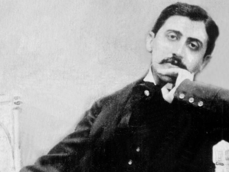 Ein schwarz-weiß Bild von Marcel Proust, der auf einem kleinen Sofa sitzt, mit schwarzen Anzug die linke Hand zur Denkerpose ans Kien gehalten. Er schaut in die Kamera und trägt einen Schnauzer.