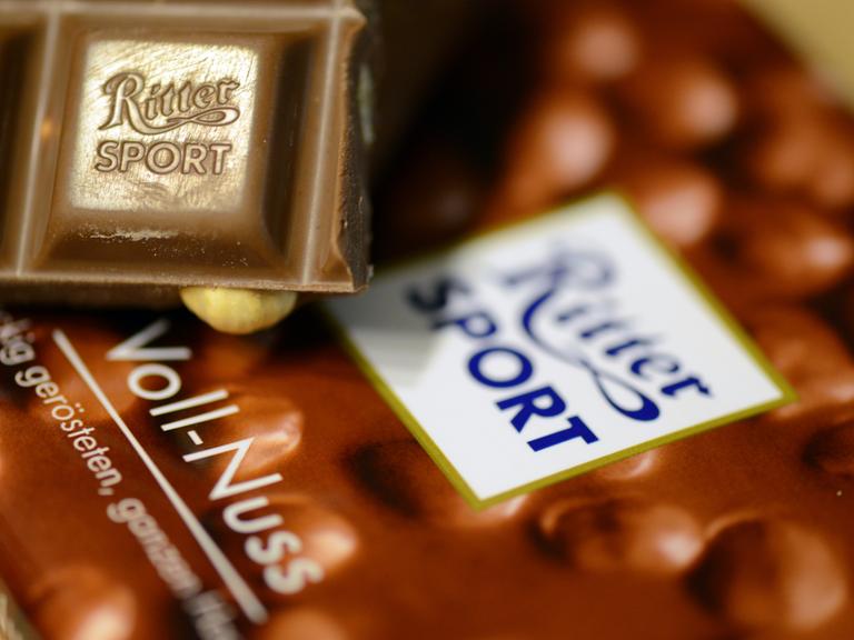 Das Bild zeigt ein kleines Stück Voll-Nuss-Schokolade der Marke Ritter-Sport. Es liegt auf einer Verpackung der Schokoladentafel.