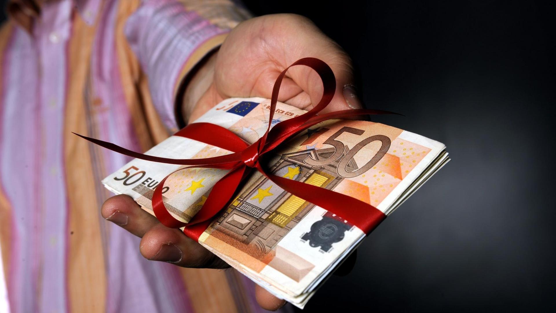 Ein Mann hält ein Bündel Geld, um das eine rote Schleife gewickelt ist. Ein Symbolbild für Korruption und Geldwäsche. Ein Mann hält ein Bündel Geld, um das eine rote Schleife gewickelt ist. Ein Symbolbild für Korruption und Geldwäsche.