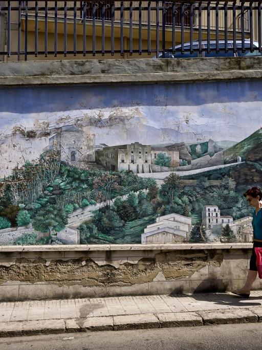 Eine Straßenstzene mit zwei älteren Passanten in Corleone, die an einem Wandbild mit dem Motiv einer ländlichen Landschat vorbeigehen.