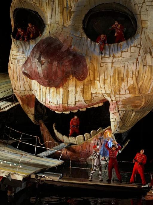 Das Foto zeigt das Bühnenbild aus "Rigoletto", inszeniert von Philipp Stölzl auf der Seebühne Bregenz: Ein riesiger Holzkopf, um den sich die Sänger gruppieren.