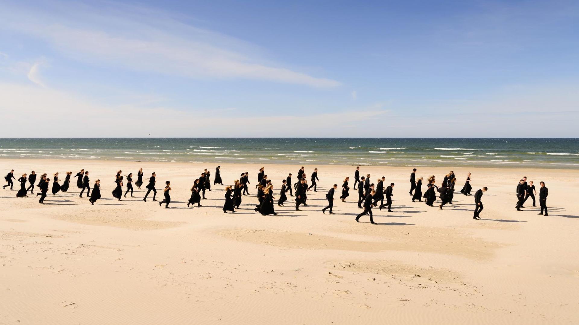 Die Orchestermitglieder laufen in schwarzer Kleidung einen langen Strand entlang. Die Orchestermitglieder laufen in schwarzer Kleidung einen langen Strand entlang.