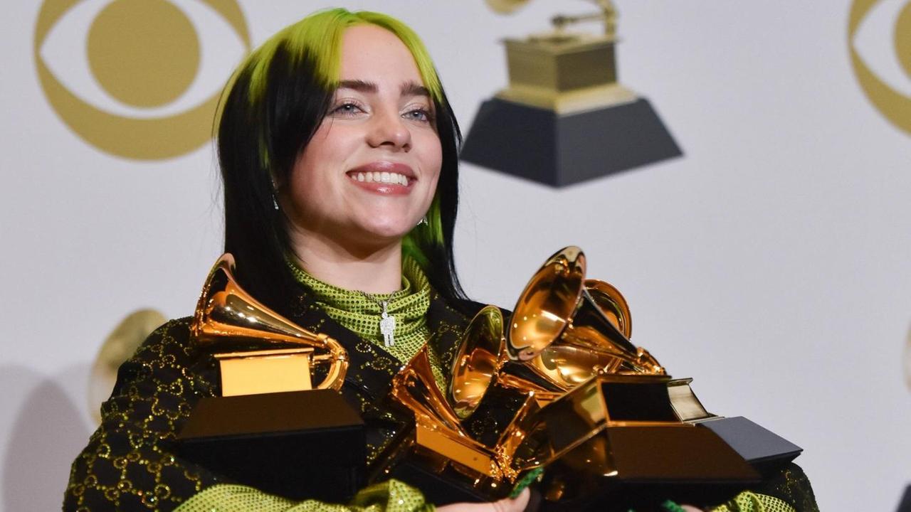 Die Sängerin Billie Eilish hat fünf goldene Preise in der Hand. Sie lacht. Sie hat grün-schwarz gefärbte Haare und trägt schwarze Kleidung. Die Sängerin Billie Eilish hat fünf goldene Preise in der Hand. Sie lacht. Sie hat grün-schwarz gefärbte Haare und trägt schwarze Kleidung.