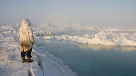 Ein Inuit in traditioneller Fellkleidung steht auf brüchigem Meereis und schaut in die Ferne.