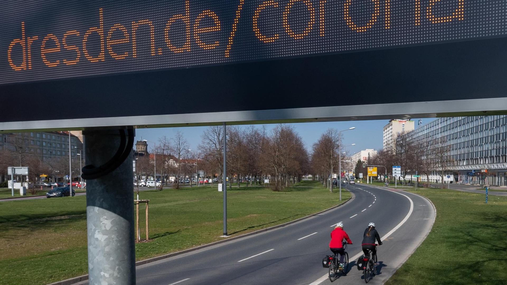 Auf einer digitalen Anzeigetafel in der Dresdner Innenstadt ist "dresden.de/corona" zu lesen während zwei Fahrradfahrerinnen auf dem Radweg vorbei fahrenung weltweit