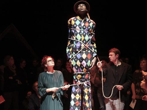 Szenenfoto: Ein bunt gekleideter Mann mit dunkler Hautfarbe steht auf einem Podest. Eine weiße Frau und ein weißer Mann haben ein Seil mehrfach um seinen Körper gespannt. Drum herum und im Hintergrund sind Menschen.
