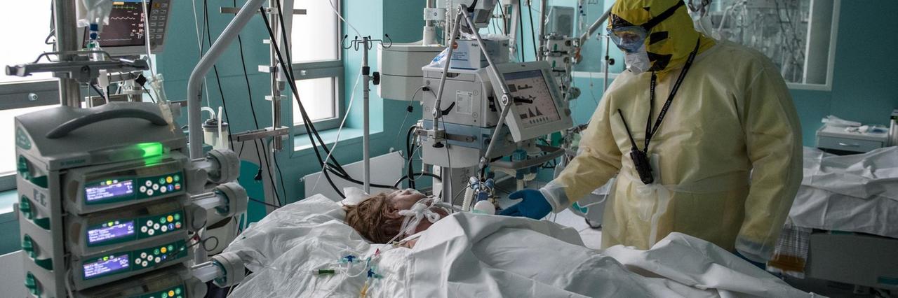 Der Arzt in voller Schutzkleidung steht an der Bettseite der Patientin, die unter einer weißen Decke liegt. Um sie herum Geräte, Kabel und Schläuche. Der Arzt in voller Schutzkleidung steht an der Bettseite der Patientin, die unter einer weißen Decke liegt. Um sie herum Geräte, Kabel und Schläuche.