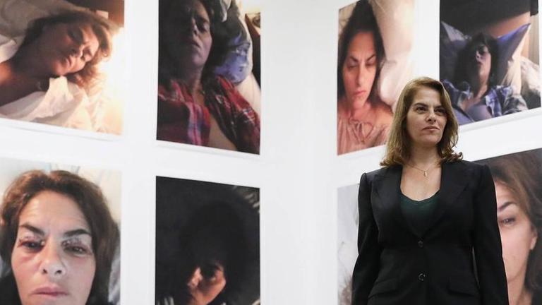 Die Künstlerin in einem hellen Raum. An den Wänden sind Selfies von ihr zu sehen, die die Künstlerin müde und matt zeigen. Die Künstlerin in einem hellen Raum. An den Wänden sind Selfies von ihr zu sehen, die die Künstlerin müde und matt zeigen.