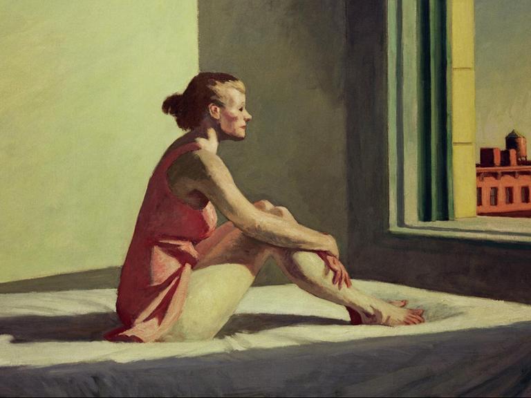 Eine junge Frau sitzt auf einem Bett und blickt melancholisch aus dem Fenster.