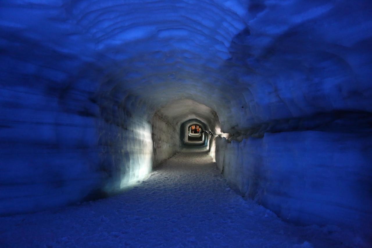 Das Eis des Tunnelgangs ist im Vordergrund in dunklem Blau angeleuchtet. Weiter hinten wird es weiß, ganz hintern ist eine Gruppe von menschen zu sehen. Das Eis des Tunnelgangs ist im Vordergrund in dunklem Blau angeleuchtet. Weiter hinten wird es weiß, ganz hintern ist eine Gruppe von menschen zu sehen.
