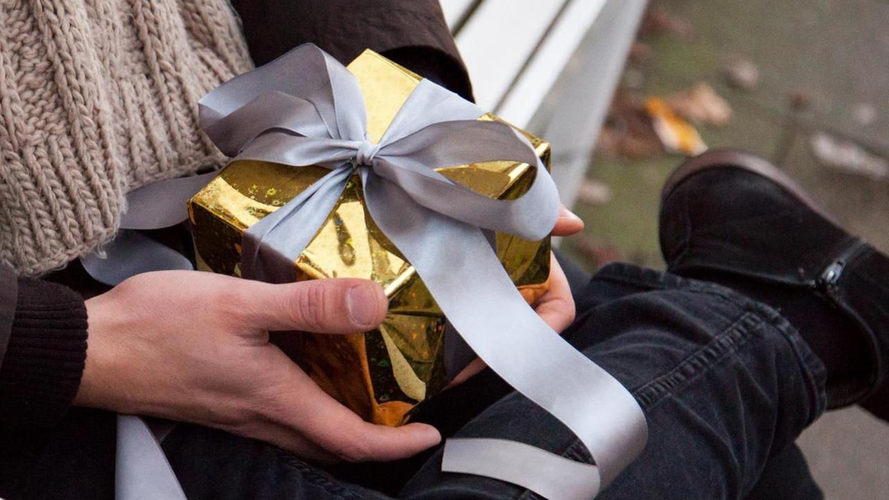 Ein junger Mann sitzt auf einer Bank und hält ein kleines, in goldenes Papier verpacktes, Geschenk in der Hand. Ein junger Mann sitzt auf einer Bank und hält ein kleines, in goldenes Papier verpacktes, Geschenk in der Hand.