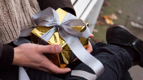 Ein junger Mann sitzt auf einer Bank und hält ein kleines, in goldenes Papier verpacktes, Geschenk in der Hand.