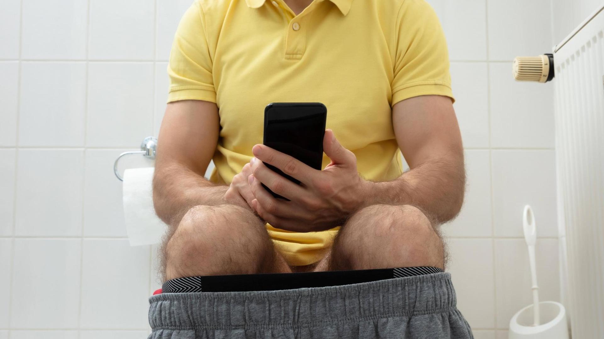 Ein Mann sitzt mit heruntergelassener Hose und Smartphone auf der Toilette. Ein Mann sitzt mit heruntergelassener Hose und Smartphone auf der Toilette.