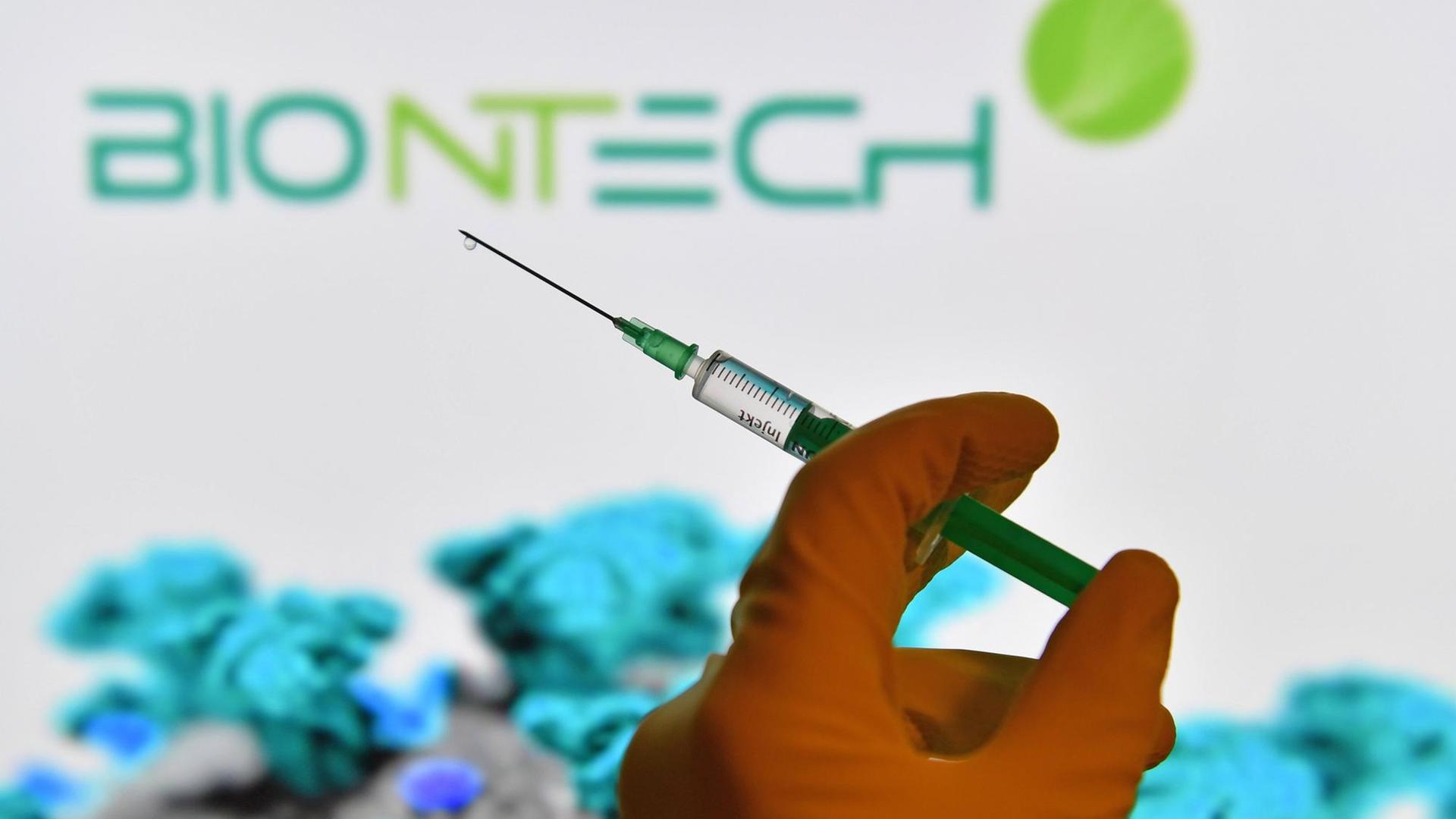 Symbolfoto: Eine Hand in Gummihandschuhen hält eine Einwegspritze mit Impfstoff zur Injektion mit einer Kanuele, im Hintergrund ist das Logo von "Biontech" zus sehen