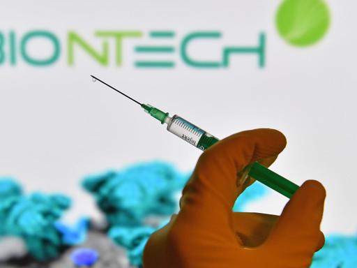 Symbolfoto: Eine Hand in Gummihandschuhen hält eine Einwegspritze mit Impfstoff zur Injektion mit einer Kanuele, im Hintergrund ist das Logo von "Biontech" zus sehen