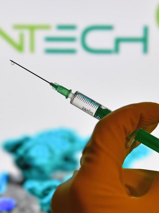 Symbolfoto: Eine Hand in Gummihandschuhen hält eine Einwegspritze mit Impfstoff zur Injektion mit einer Kanuele, im Hintergrund ist das Logo von "Biontech" zus sehen