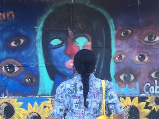 Eine junge Frau mit langem schwarzen Zopf und Fahrrad betrachtet ein Wandgemälde, das ein Frauengesicht mit leeren Augenhöhlen zeigt, drumherum viele Augen.