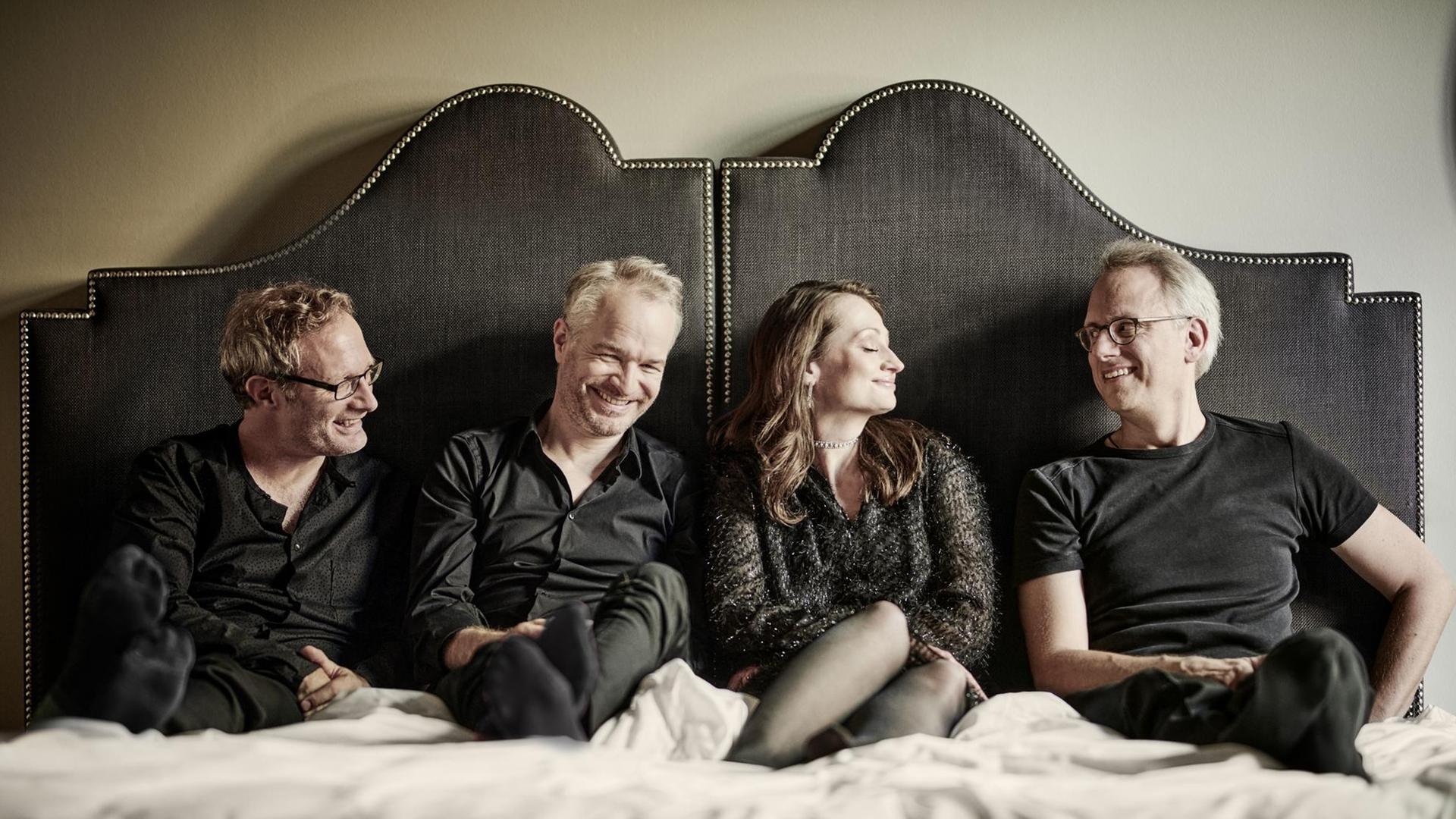 Eine Frau und drei Männer sitzen in einem Bett mit Kopfteil und lächeln einander an.