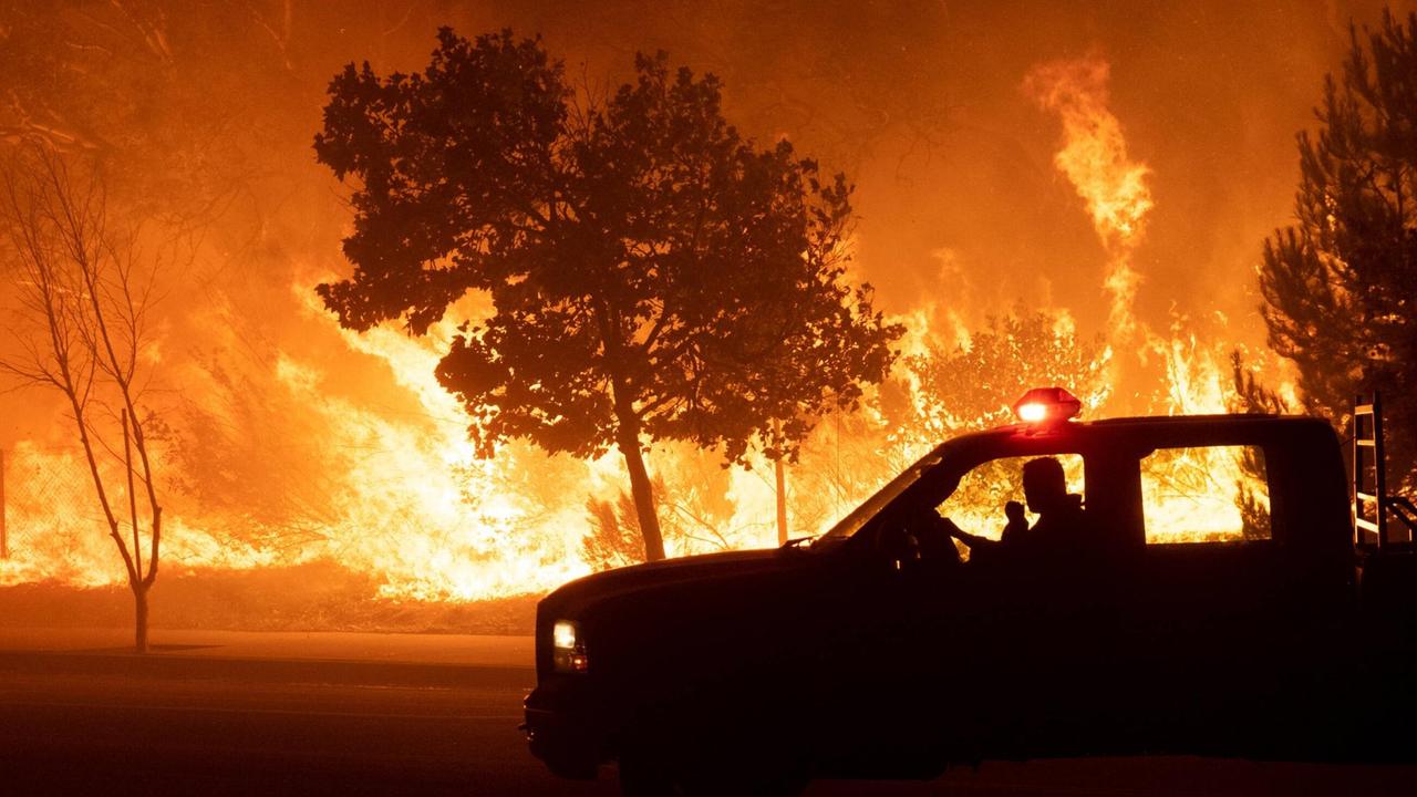 Ein Waldbrandpatrouillen-Auto vor brennenden Bäumen, dahinter schlagen Flammen hoch.
