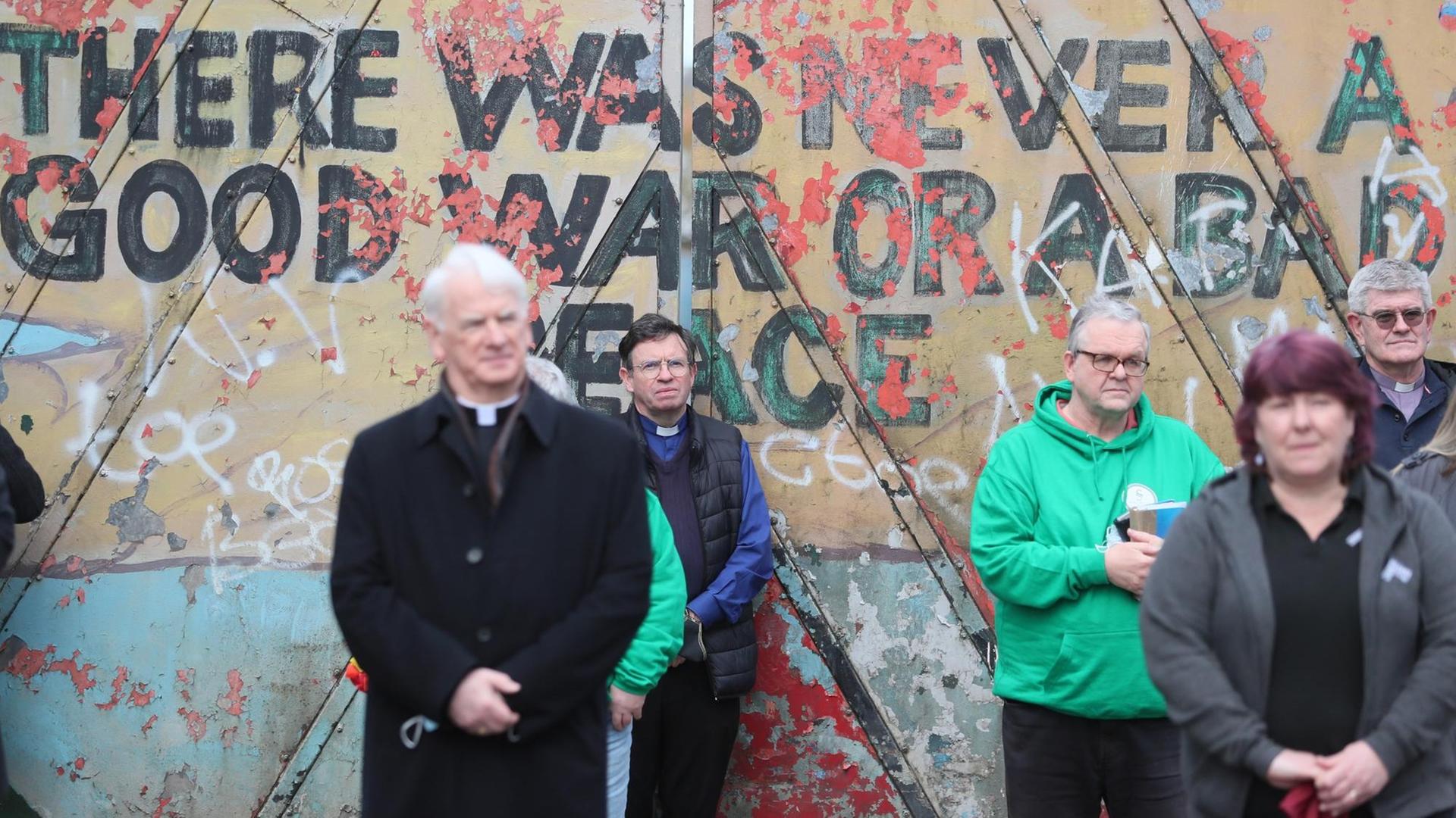 Vor einer Mauer mit dem Spruch "There was never a good war or a bad peace" stehen Menschen mit gefalteten Händen. Vor einer Mauer mit dem Spruch "There was never a good war or a bad peace" stehen Menschen mit gefalteten Händen.