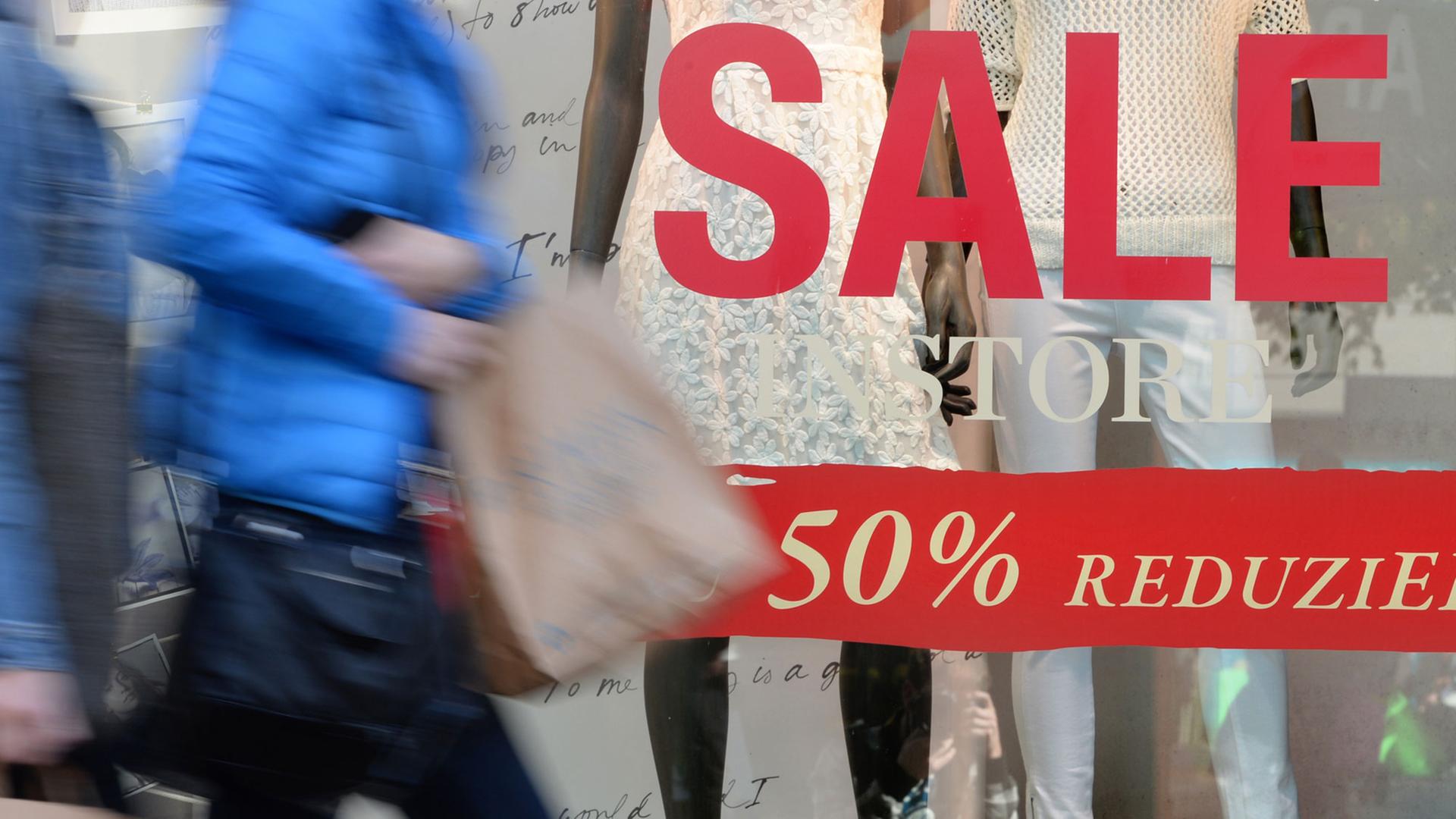 Teil eines Schaufensters mit dem Aufdruck "Sale - 50 % reduziert", davor eine vorbeigehende Frau mit Einkaufstüte