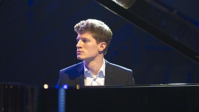 EIn junger Mann mit kurzen blonden Haaren sitzt in Jackett und Hemd gekleidet an einem geöffneten Konzertflügel und blickt zur Seite.
