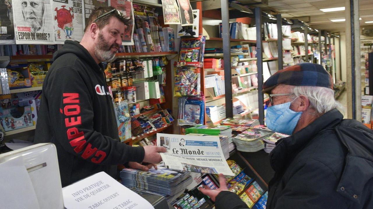 Ein Kioskbesitzer in Paris verkauft einem Kunden mit Mundschutz eine Tageszeitung Ein Kioskbesitzer in Paris verkauft einem Kunden mit Mundschutz eine Tageszeitung