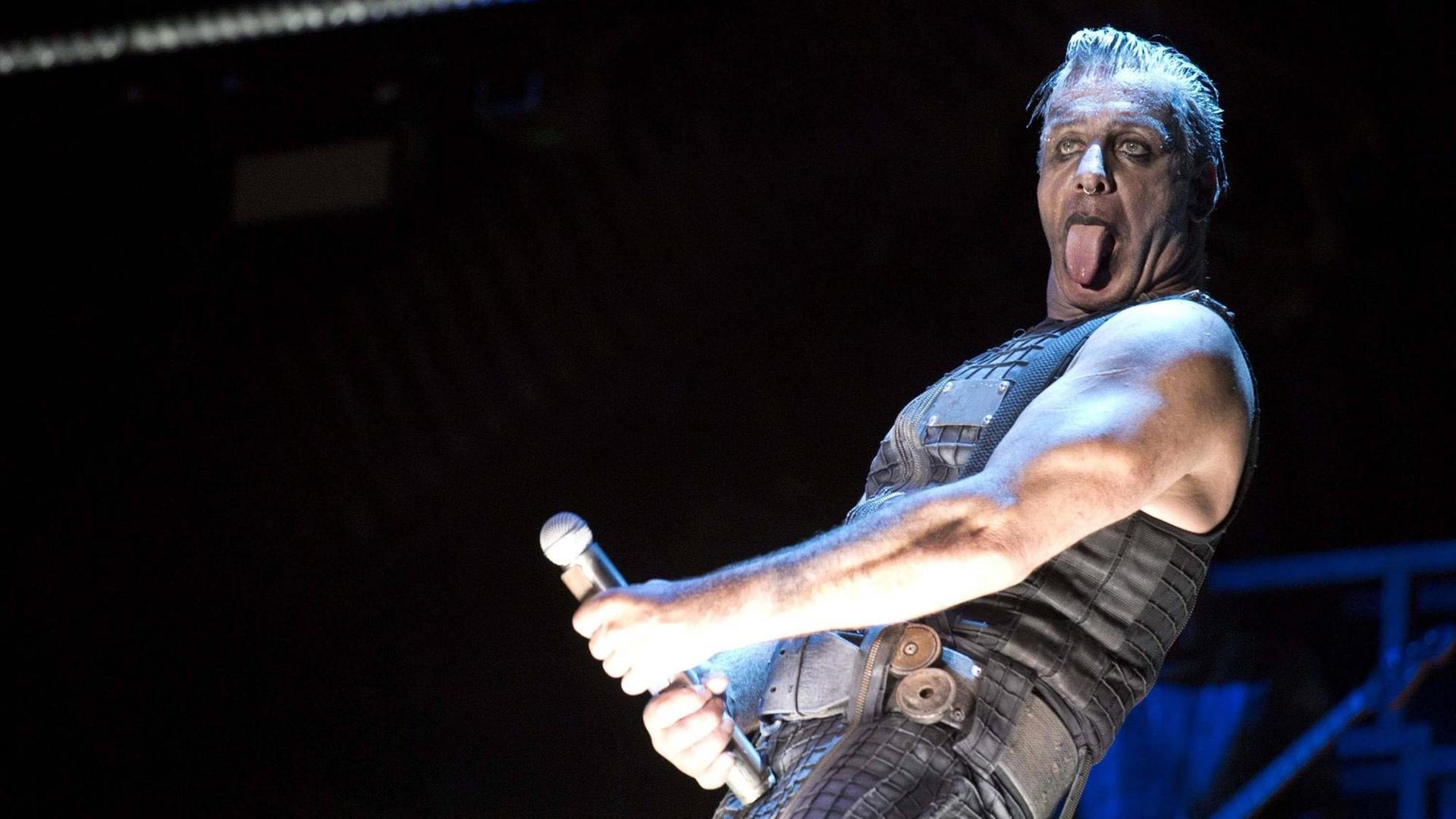 Der Sänger der Band Rammstein steht auf einer dunklen Bühne und streckt seine Zunge raus. Das Mikro hält er in der rechten Hand in frivoler Geste vor seinen Unterleib.