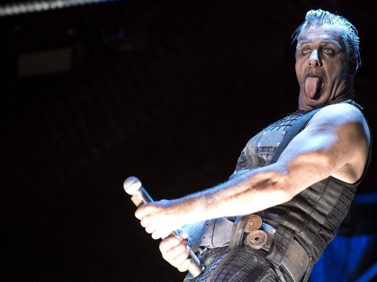 Der Sänger der Band Rammstein steht auf einer dunklen Bühne und streckt seine Zunge raus. Das Mikro hält er in der rechten Hand in frivoler Geste vor seinen Unterleib.