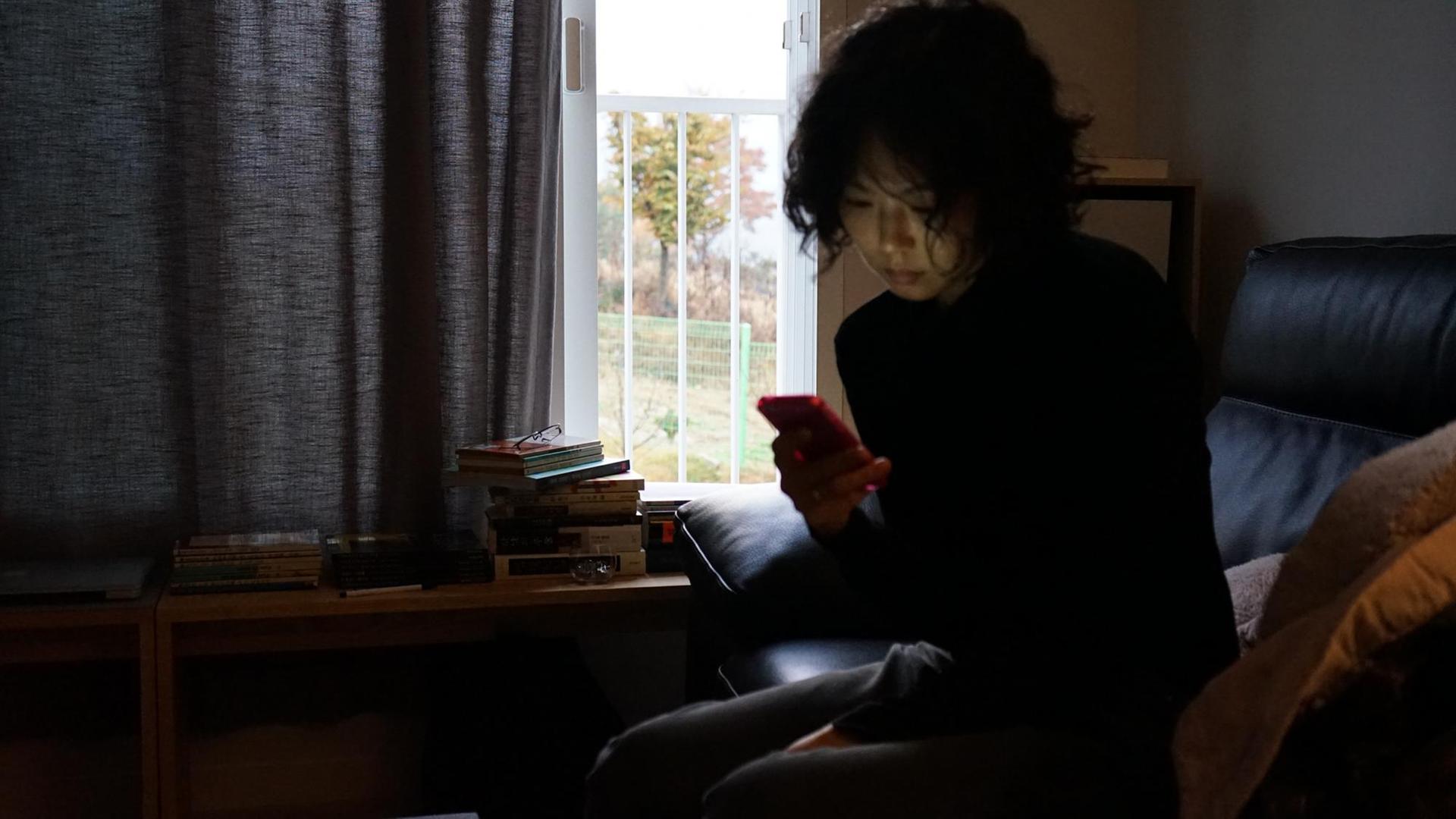 Filmstill aus "The Woman Who Ran": Eine Frau sitzt, auf das Smartphone blickend, vor einem Fenster mit halb zugezogenen Vorhängen. Filmstill aus "The Woman Who Ran": Eine Frau sitzt, auf das Smartphone blickend, vor einem Fenster mit halb zugezogenen Vorhängen.