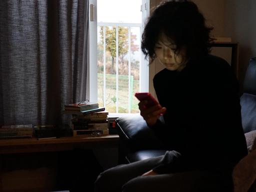 Filmstill aus "The Woman Who Ran": Eine Frau sitzt, auf das Smartphone blickend, vor einem Fenster mit halb zugezogenen Vorhängen.
